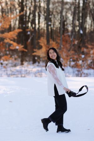 Person in a white sweater poses playfully in a snowy winter landscape with orange autumn leaves in the background.