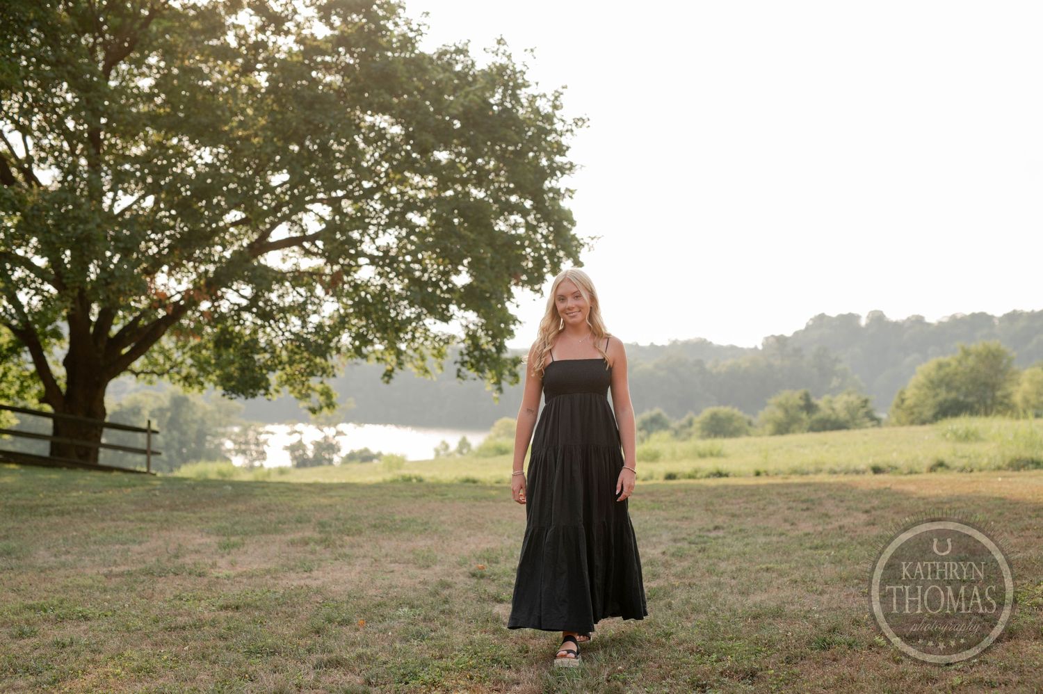 Black dress stands in golden sunlit field with trees in background.