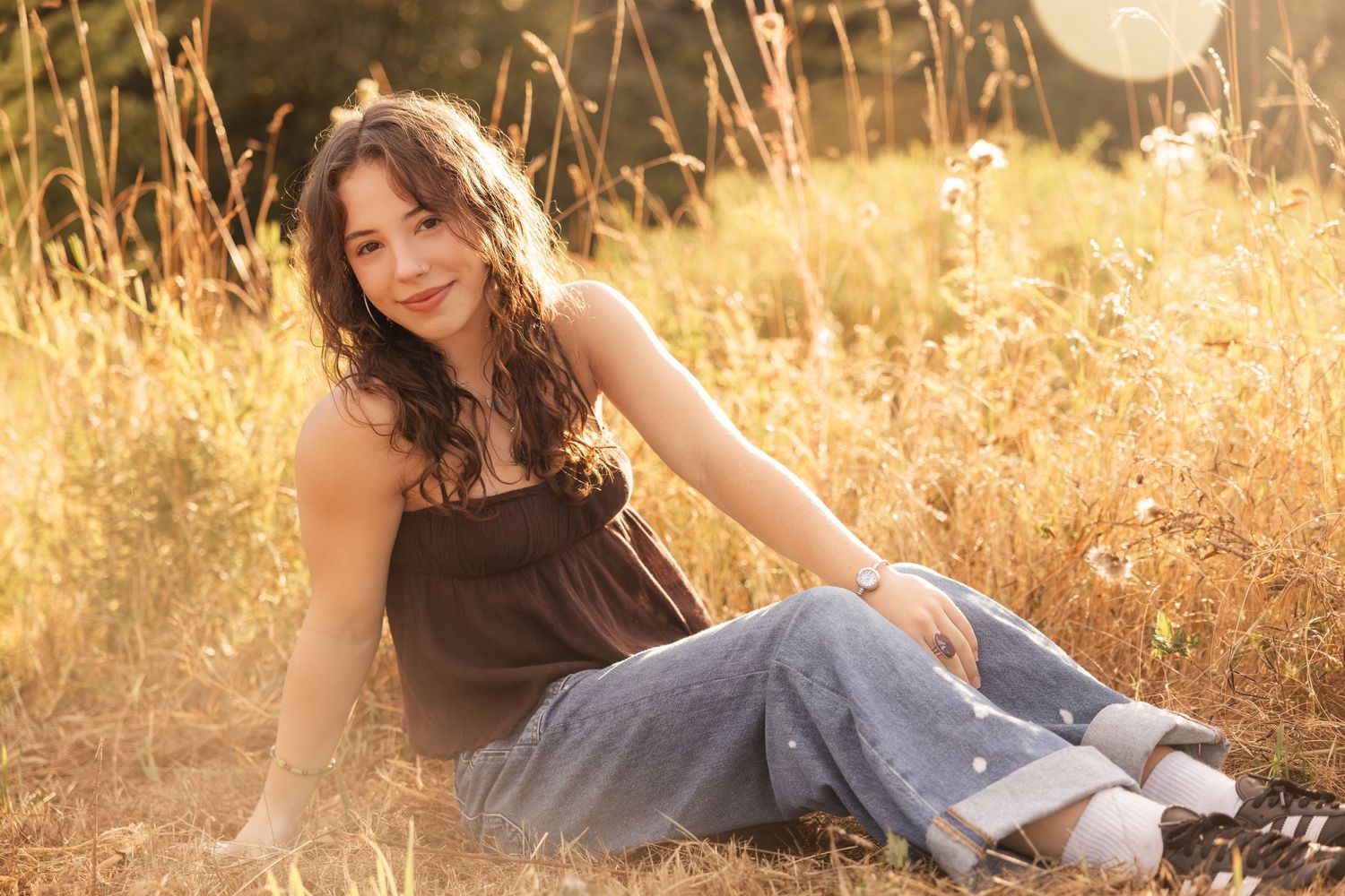 A person in a brown top and jeans sits gracefully in a sun-drenched field of golden grass at sunset.