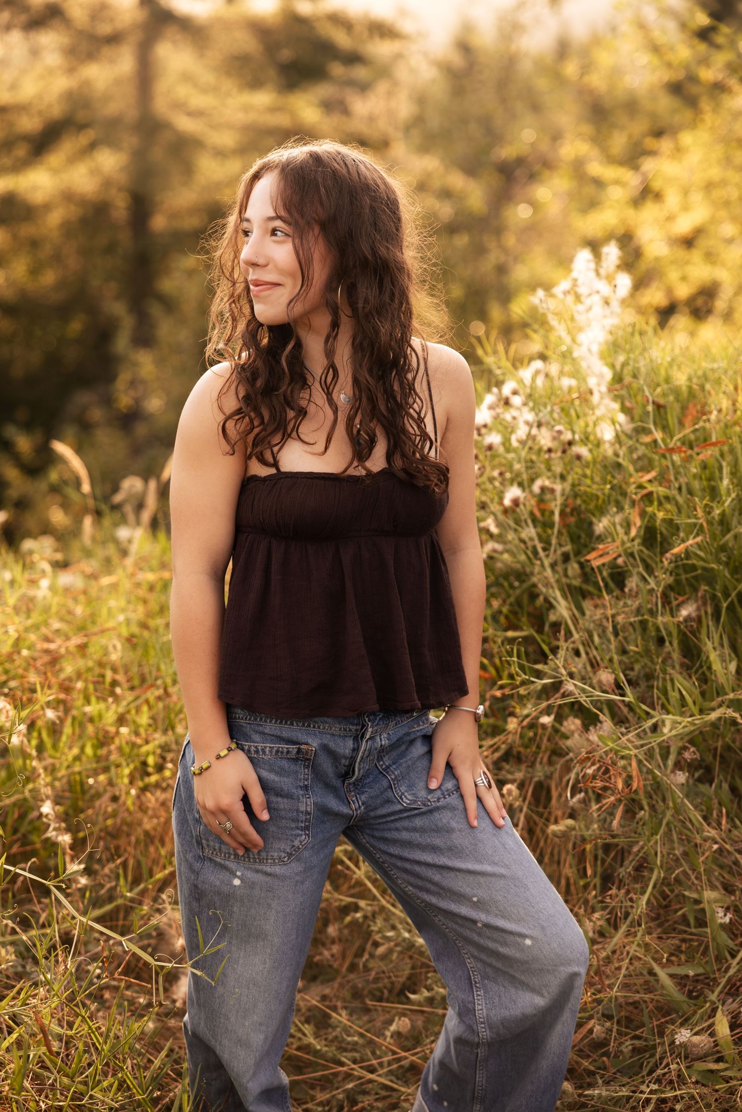 A casual outdoor portrait in a brown top and jeans amid golden sunlit wildflowers and nature.