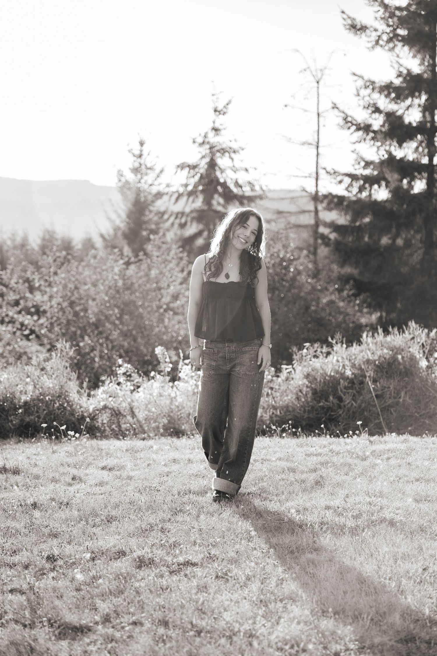 Black and white portrait of a person walking along a grassy path with evergreen trees and mountains in the background.