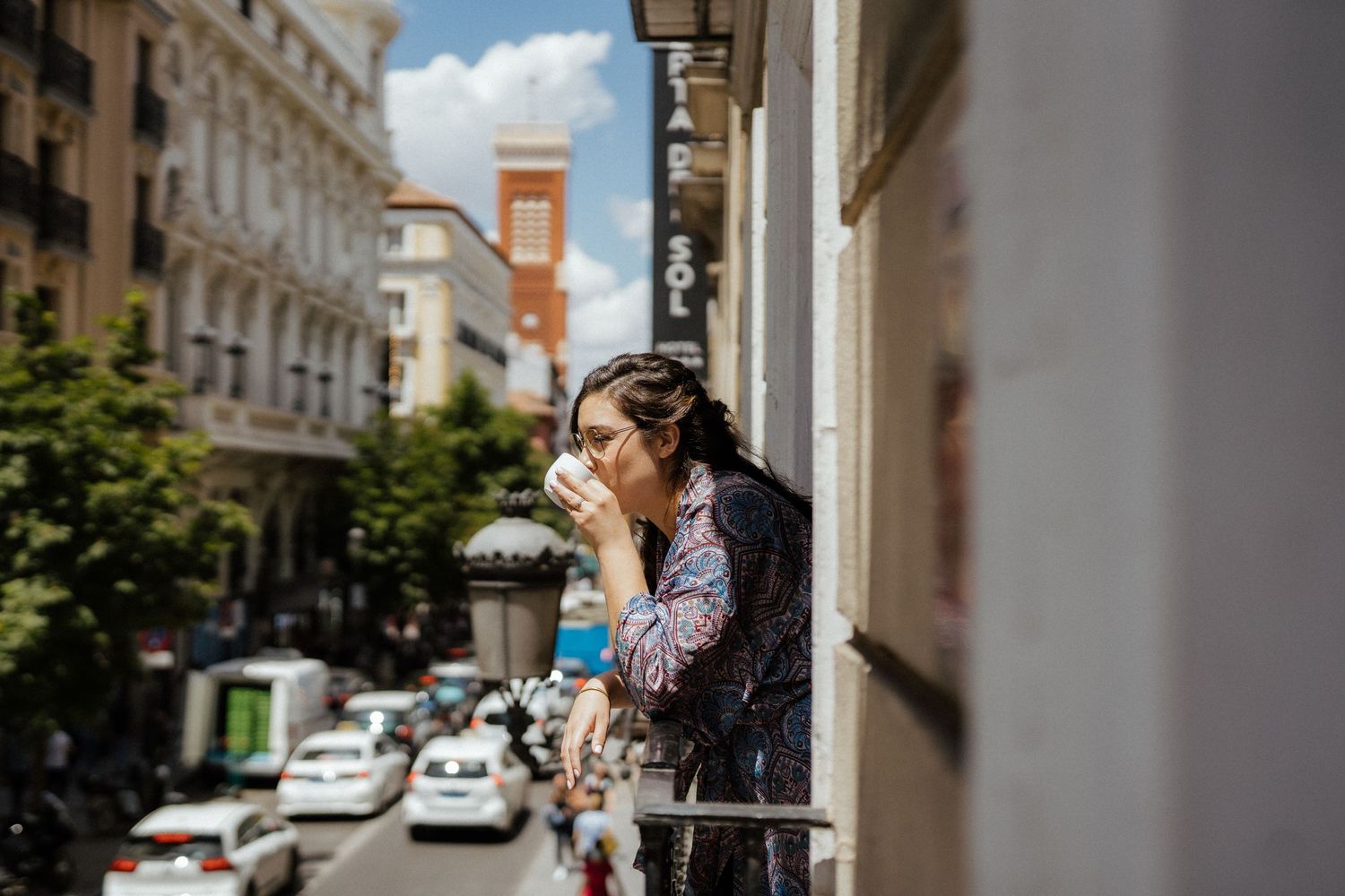 Someone standing on a balcony overlooking a sunny European city street with historic buildings and cars below.