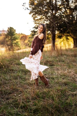 Young woman in floral skirt, brown sweater, and cowboy boots twirling in a golden field at sunset.