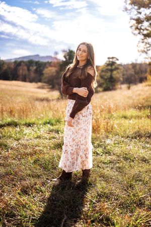 Young woman in floral skirt and dark sweater poses in sunlit autumn field with mountains in background.