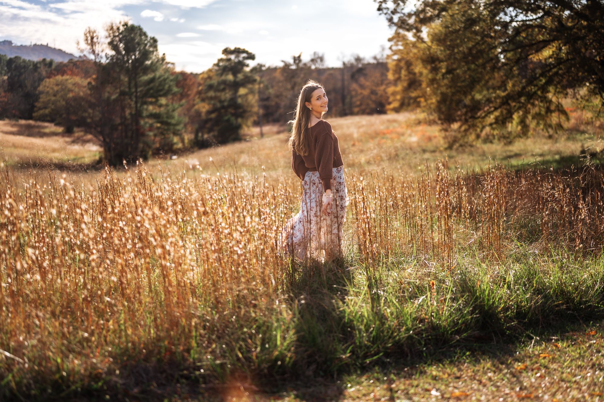 Young woman in floral skirt standing in golden autumn meadow surrounded by tall grass and colorful fall foliage.