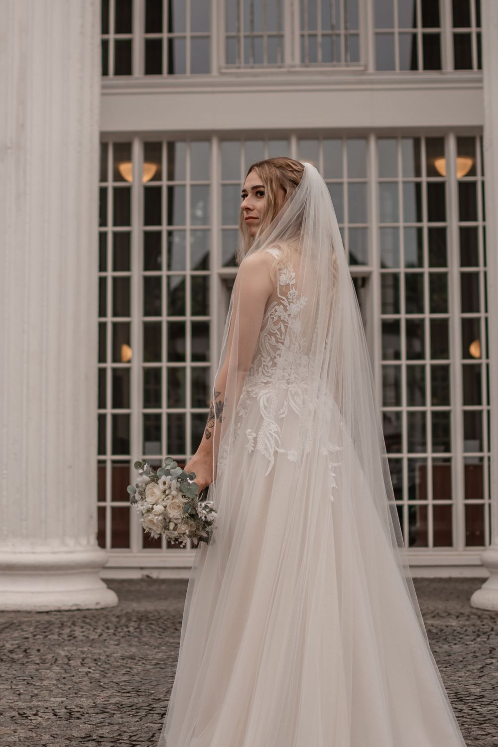 A bride in a lace wedding gown poses beside white columns with a flowing veil.