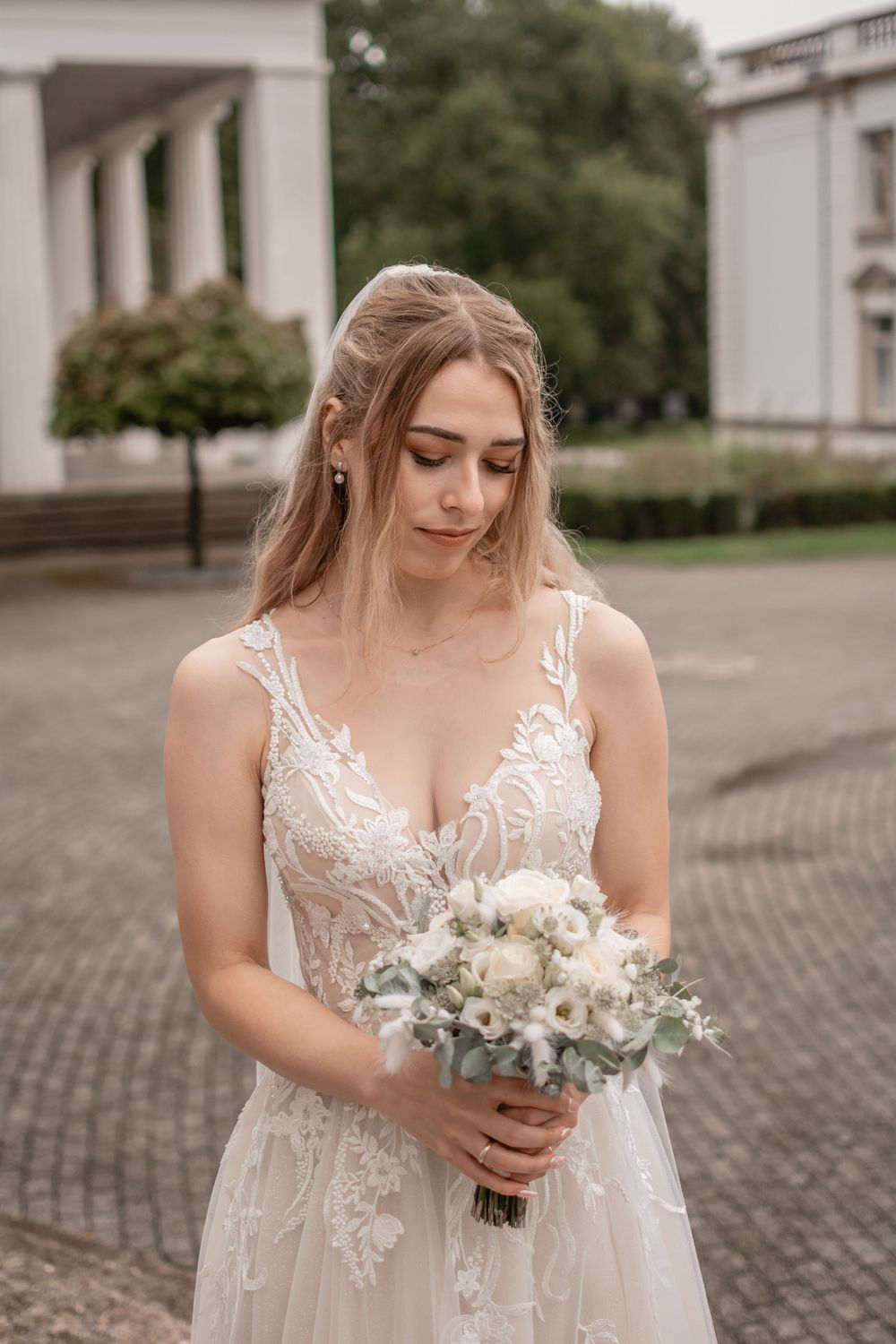 A bride in delicate lace dress holds a white bouquet against classical architecture.