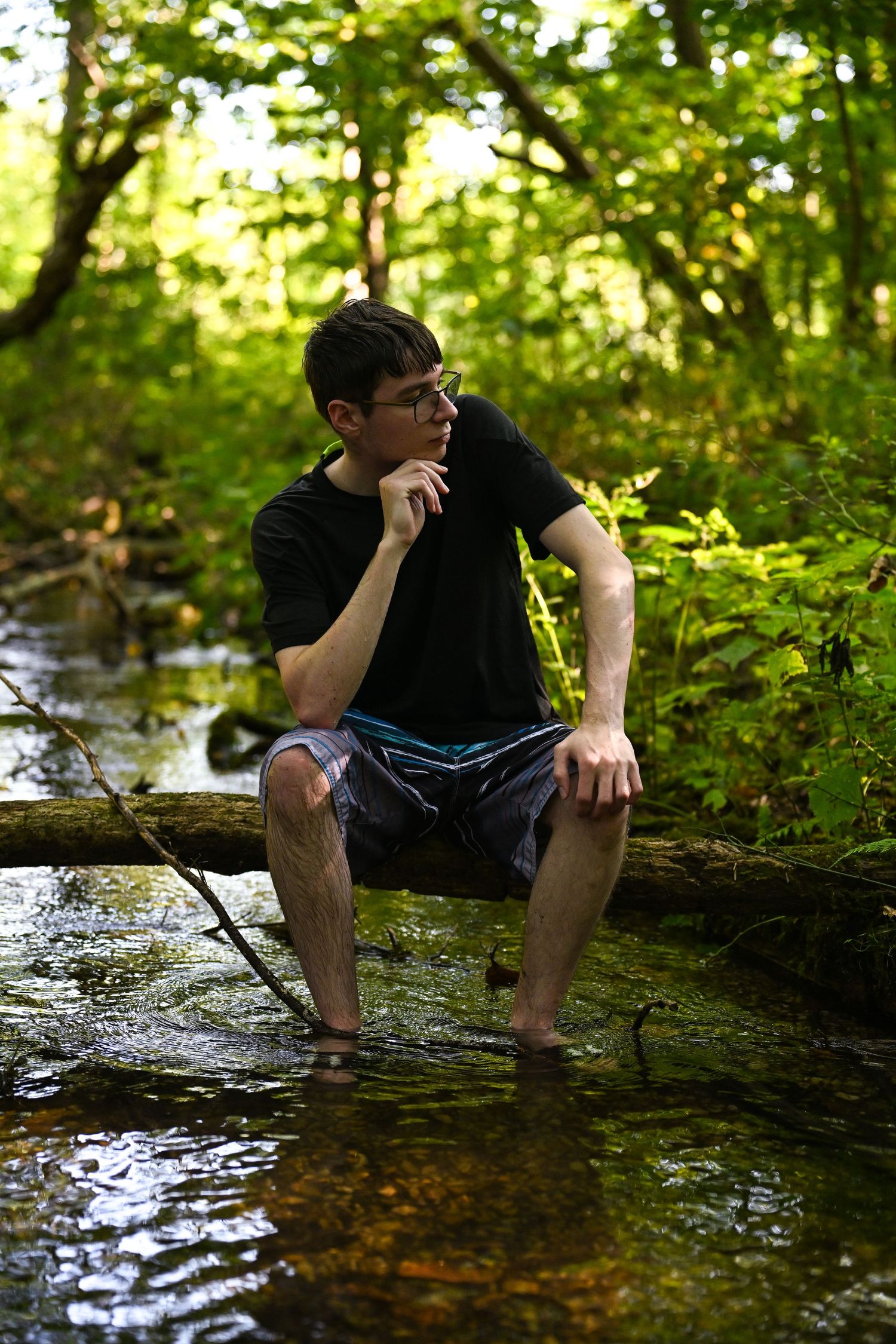 a boy with glasses and swim trunks sitting on a log in the middle of a creek