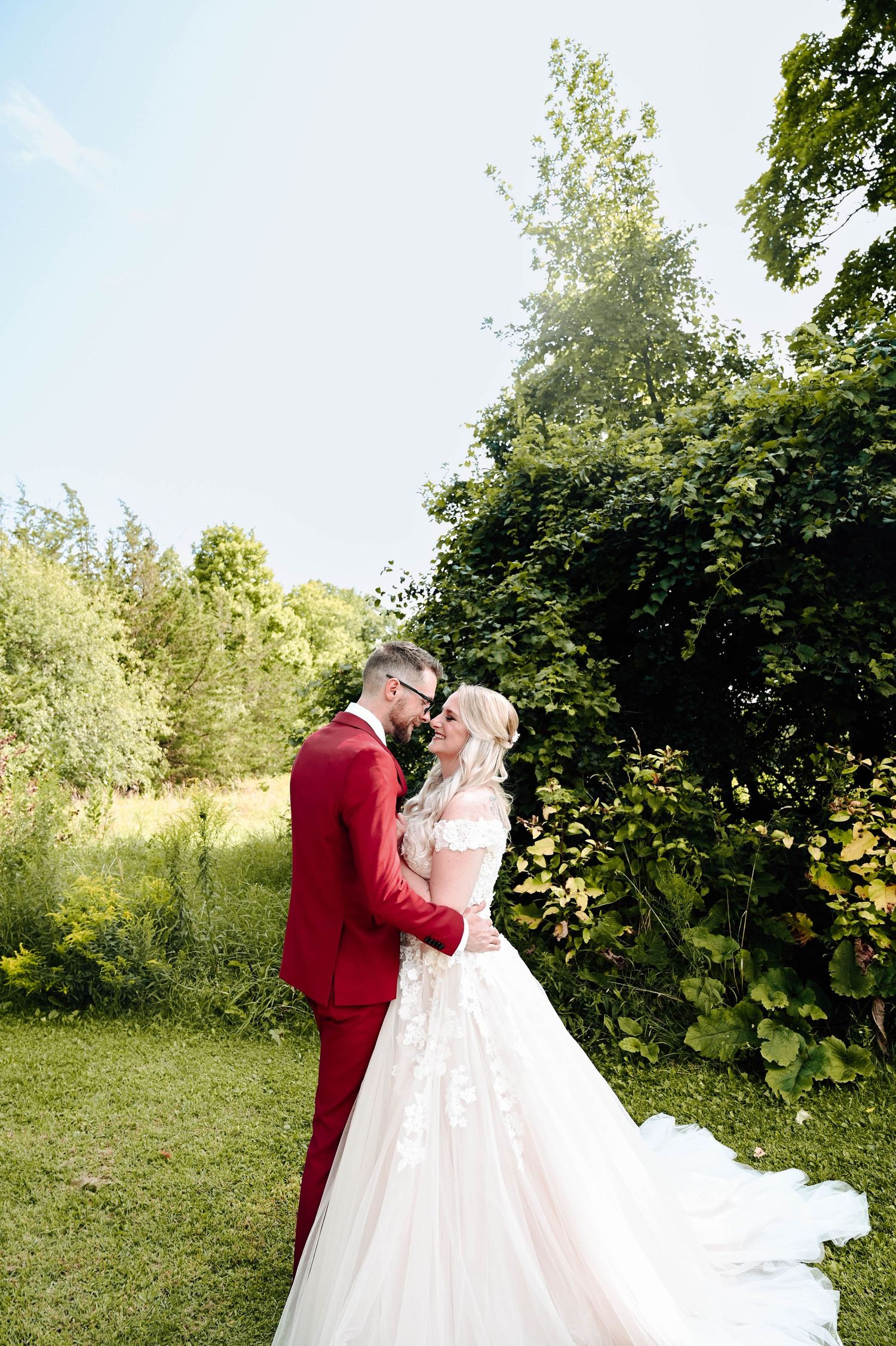 Bride in white dress and groom in red suit sharing romantic moment in garden setting.