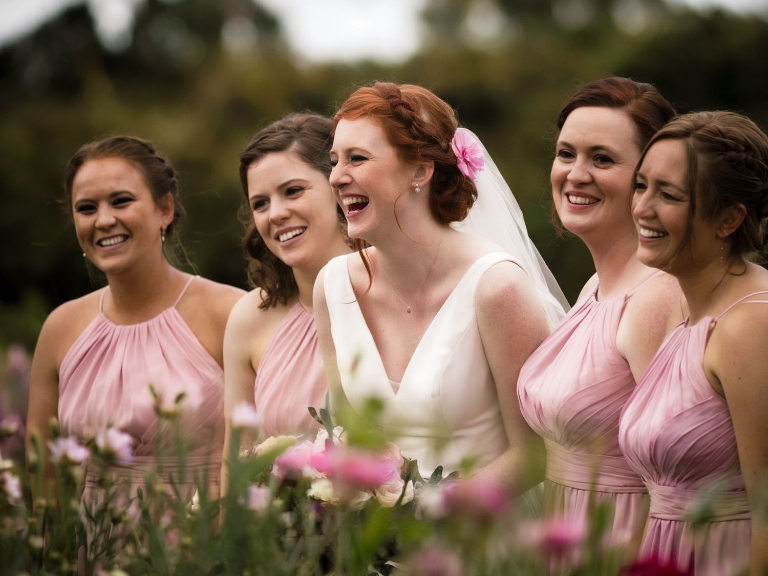 A bride in white and bridesmaids in pink dresses pose together in a wildflower meadow.