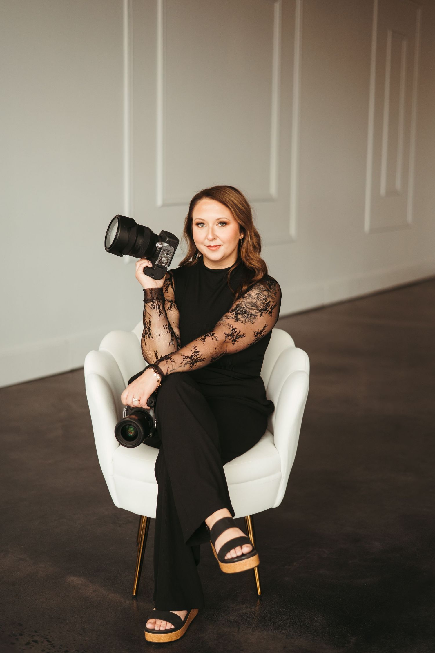 A professional female photographer poses in a white chair, holding a camera, wearing a black lace outfit.