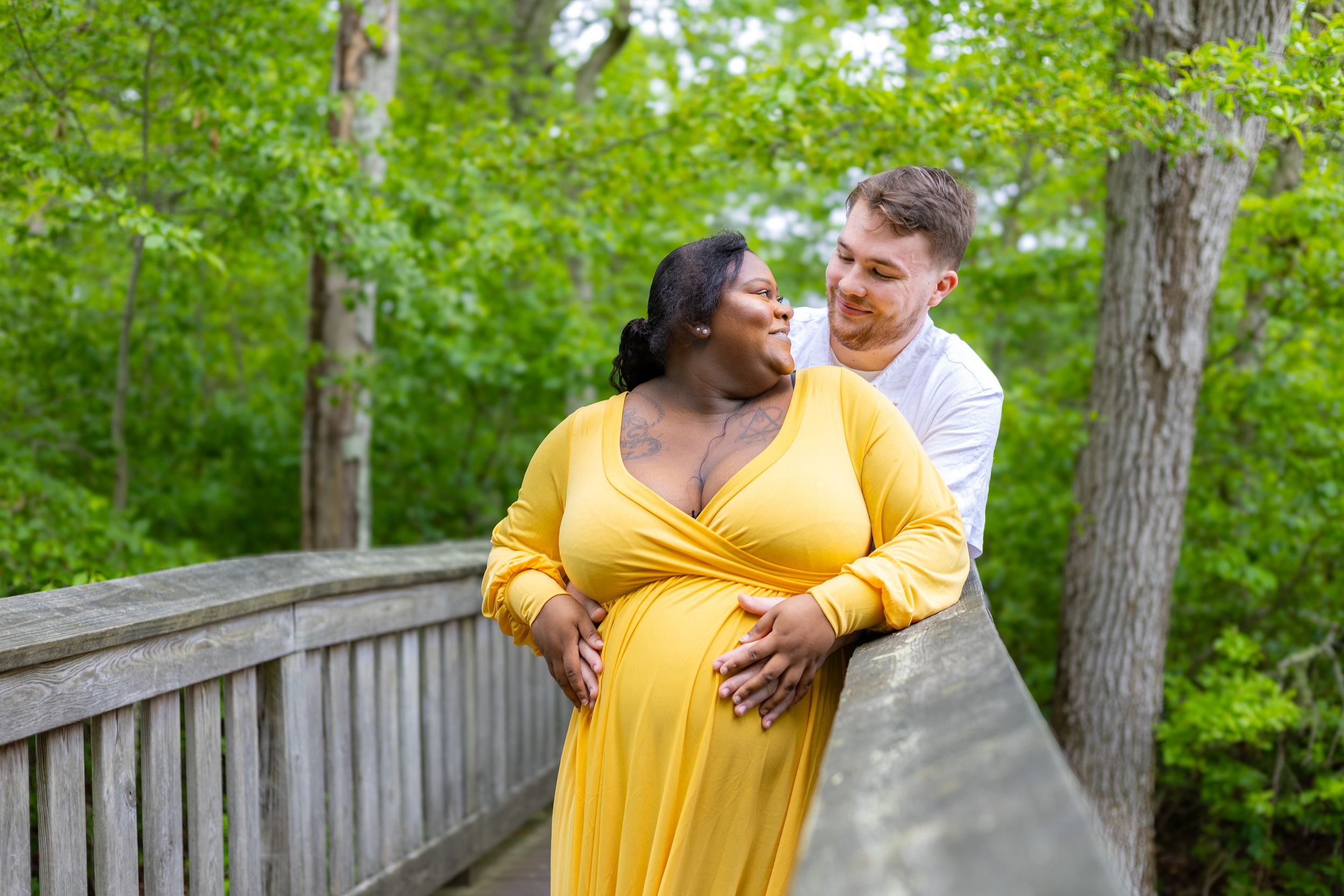 A smiling couple in a yellow dress poses for maternity photos on a wooden bridge surrounded by lush green trees on a bright day.