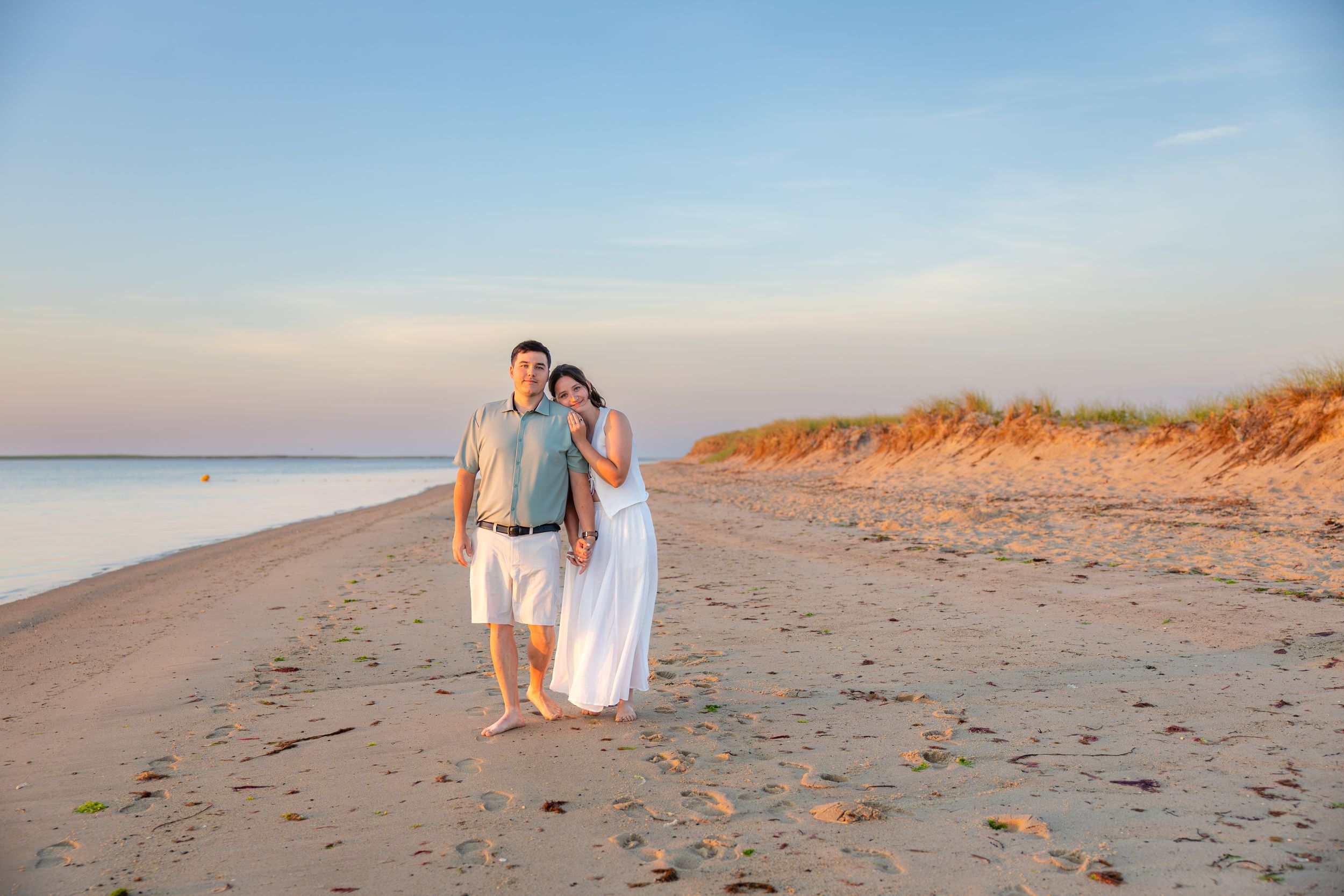 Couple embracing on a sandy beach at sunset, woman in white dress, man in gray shirt, with dunes and pastel sky.