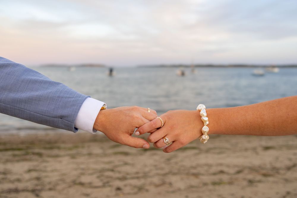 Couple holding hands on a sandy beach at sunset, woman wearing pearl bracelet and ring, man in blue suit jacket.