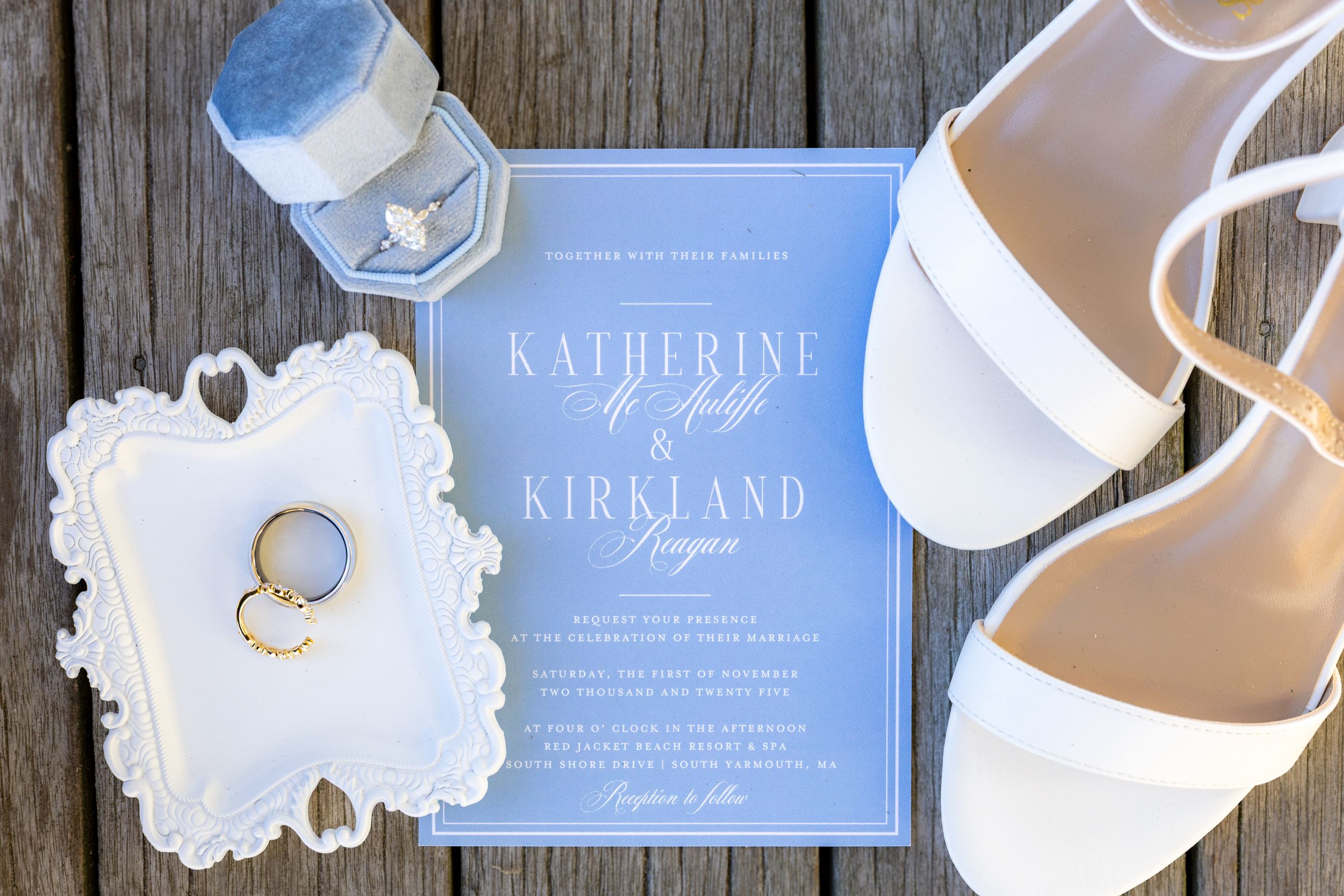 Wedding details including blue invitations, white shoes, rings, and floral accessories arranged on a rustic wooden surface.