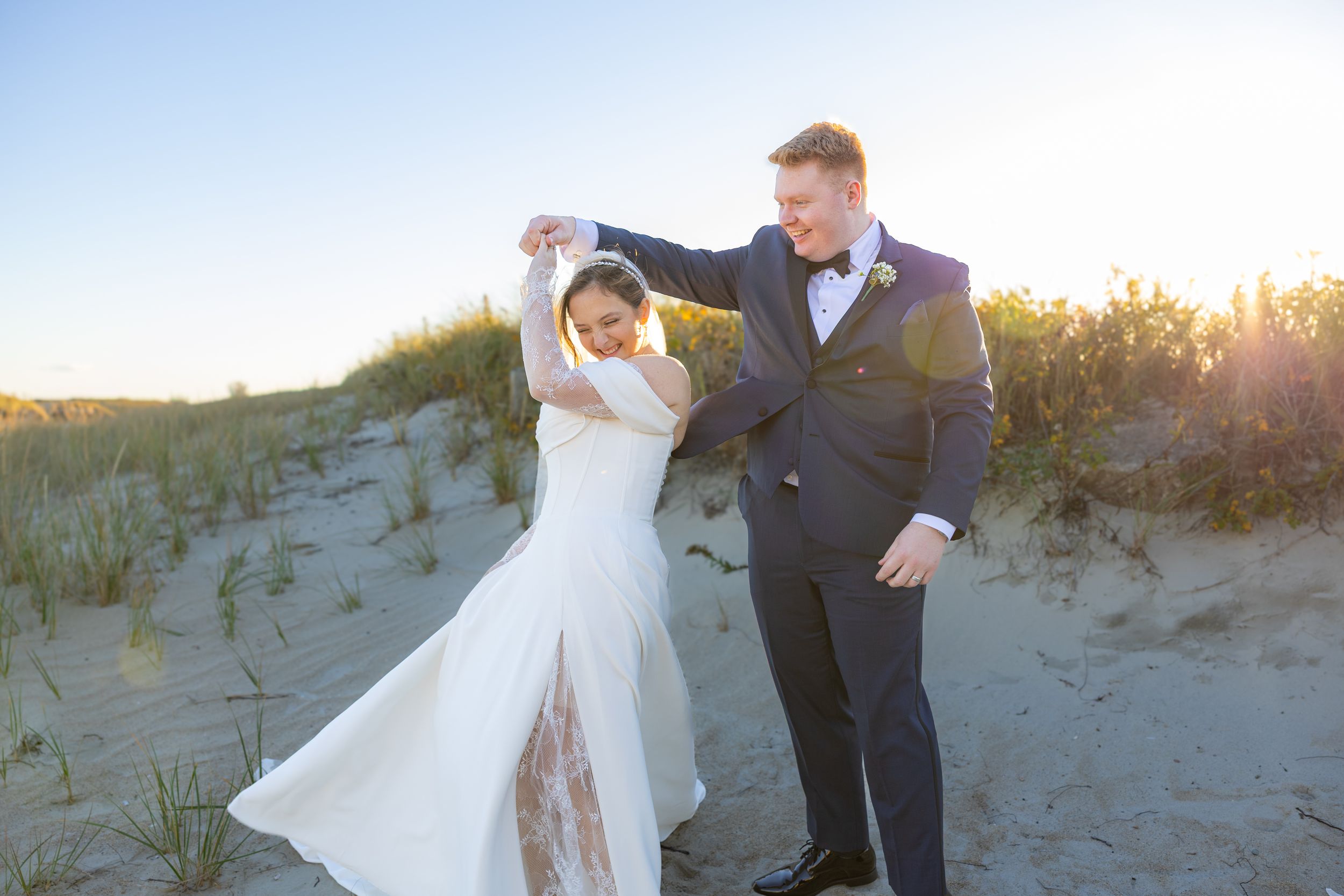 A couple dances together on sandy dunes at sunset, with the bride wearing a flowing white dress and groom in dark suit.