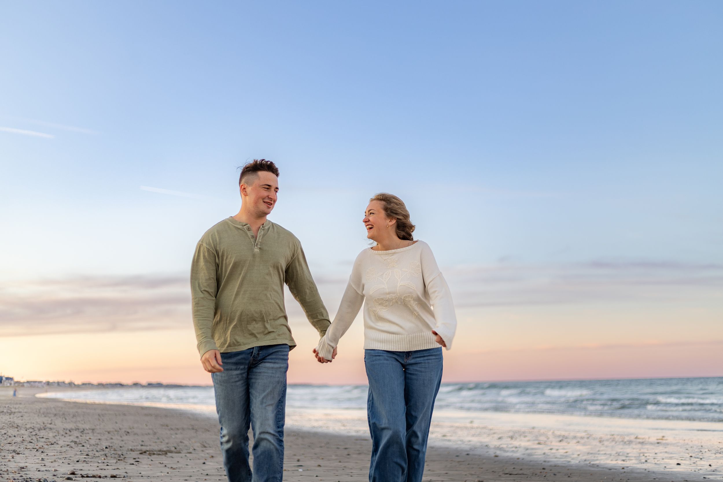 Couple holding hands walking on beach at sunset, wearing casual sweaters and jeans, smiling at each other.