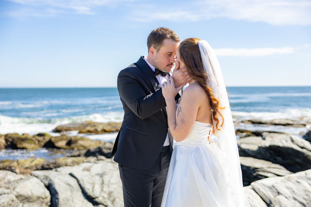 A couple shares a romantic kiss on rocky coastal cliffs with ocean waves and blue sky in the background.