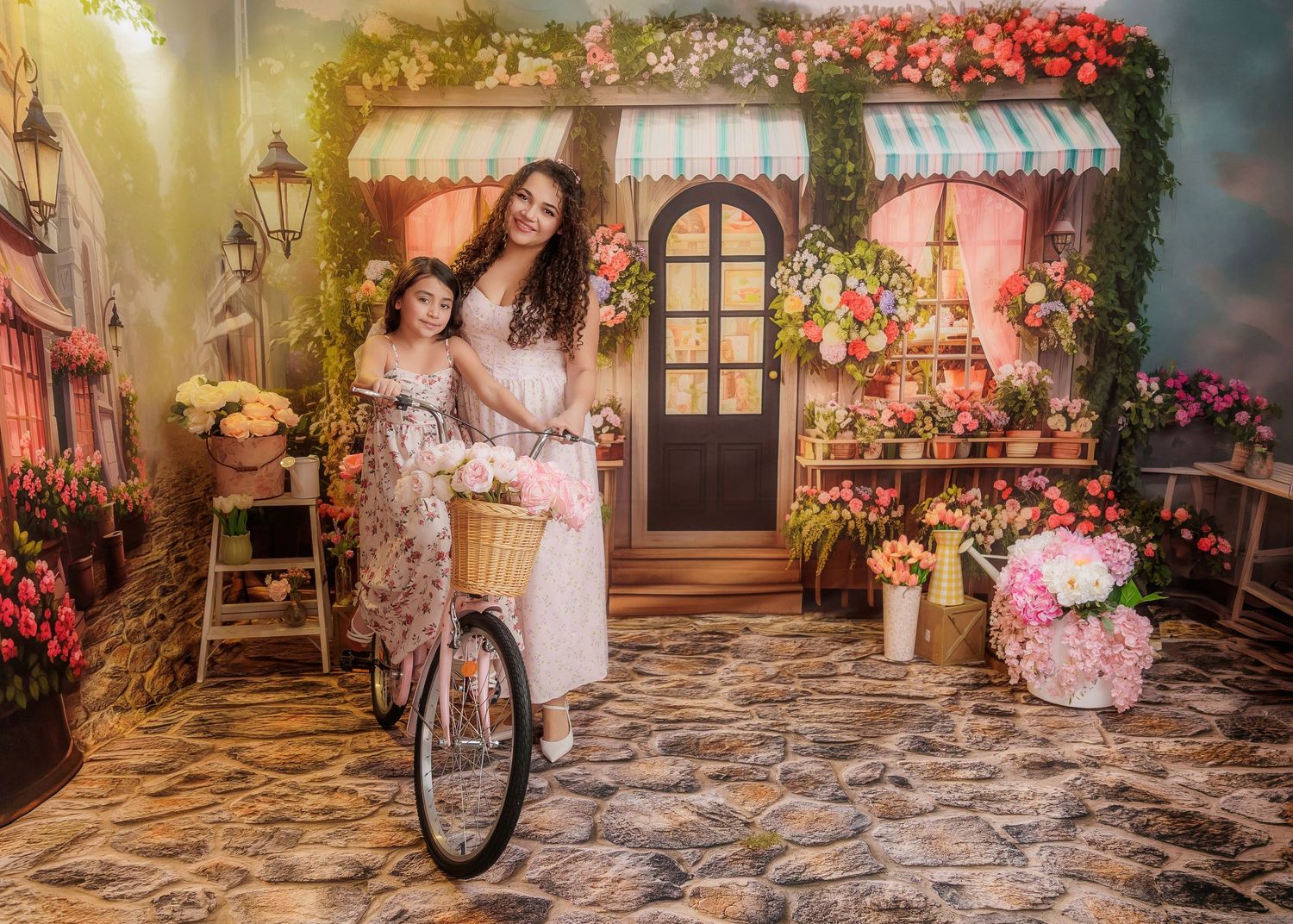 Mother and daughter in white dresses pose on a bicycle before a colorful floral shop backdrop.