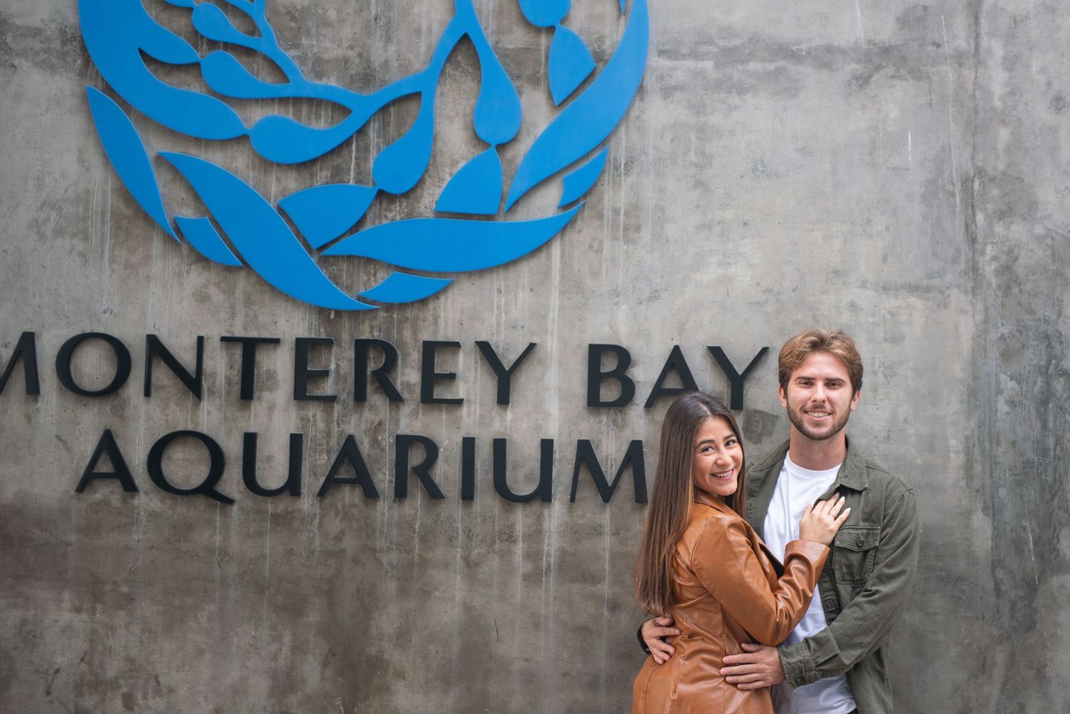 A couple poses together in front of the Monterey Bay Aquarium sign.