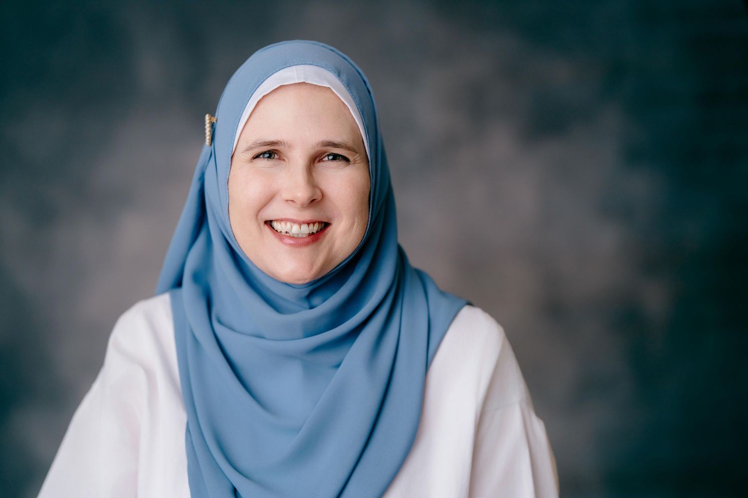 Smiling woman wearing a light blue hijab and white top against a gray studio background.