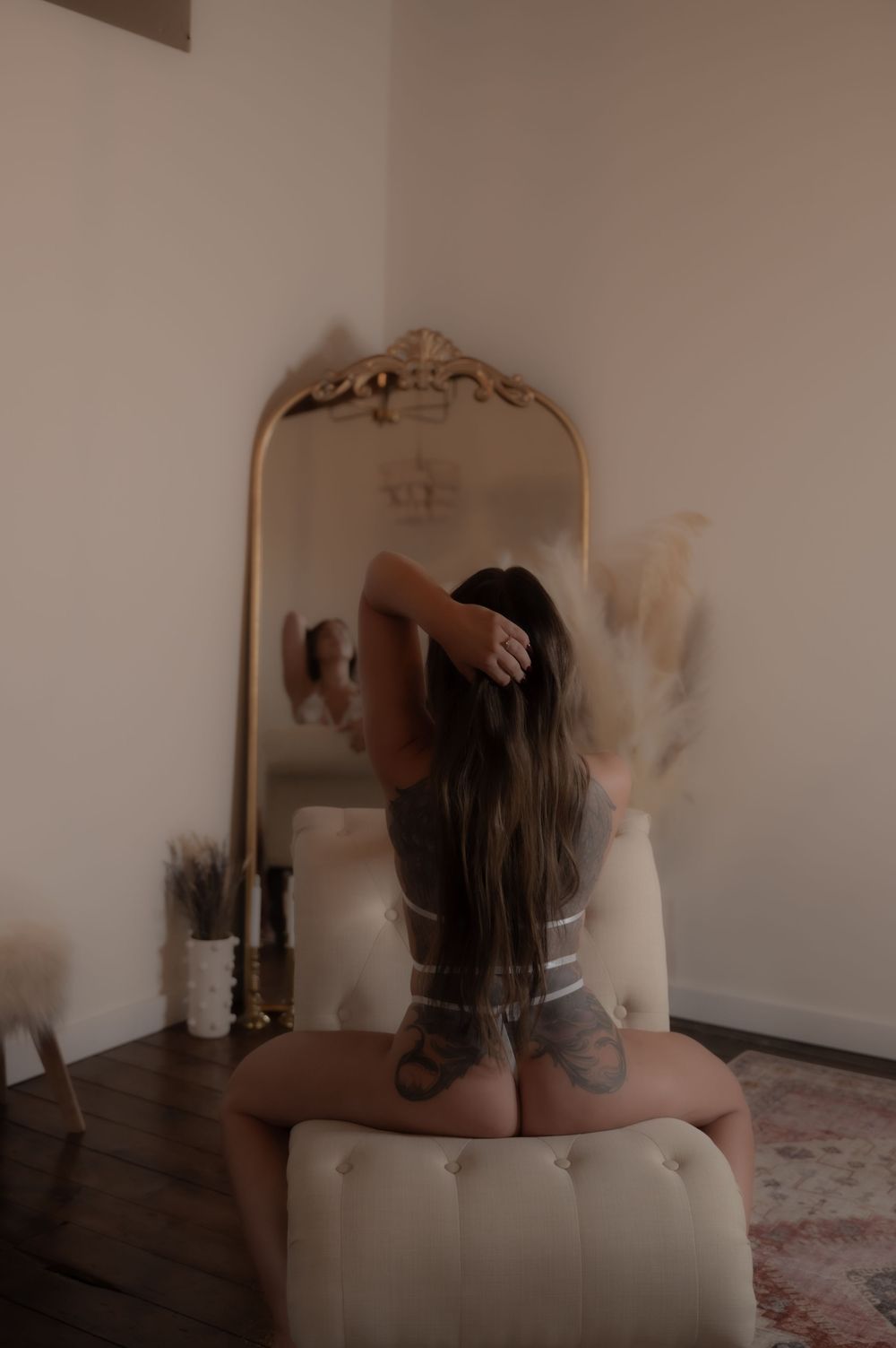 Woman with long hair and tattoos sits before ornate gold mirror in boho room with pampas grass decor.