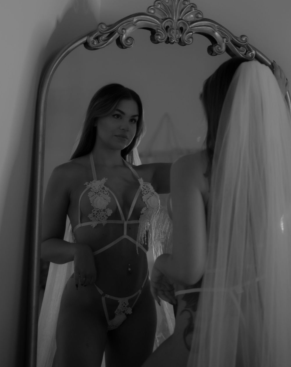 Woman in white lace lingerie and veil posing before ornate mirror in black and white photo.