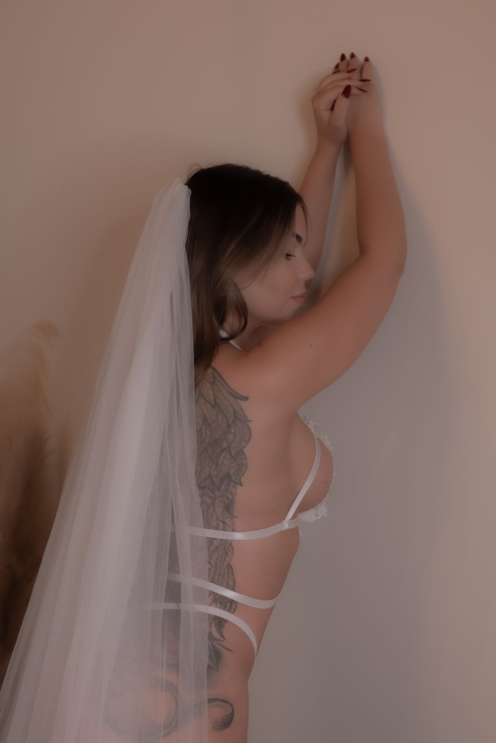 Woman in white lingerie and bridal veil with back tattoos posing against a wall.