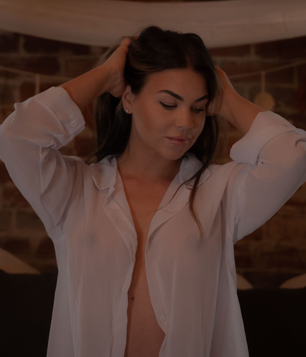 Woman in sheer white open shirt posing with hands in hair against brick wall background.