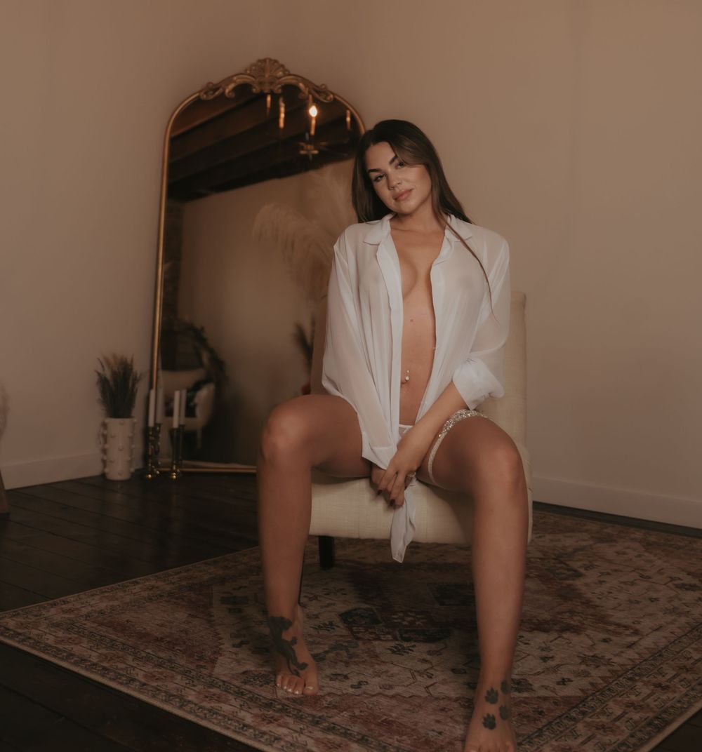 Woman in white open shirt seated on chair before gold mirror on patterned rug in warm-lit room.