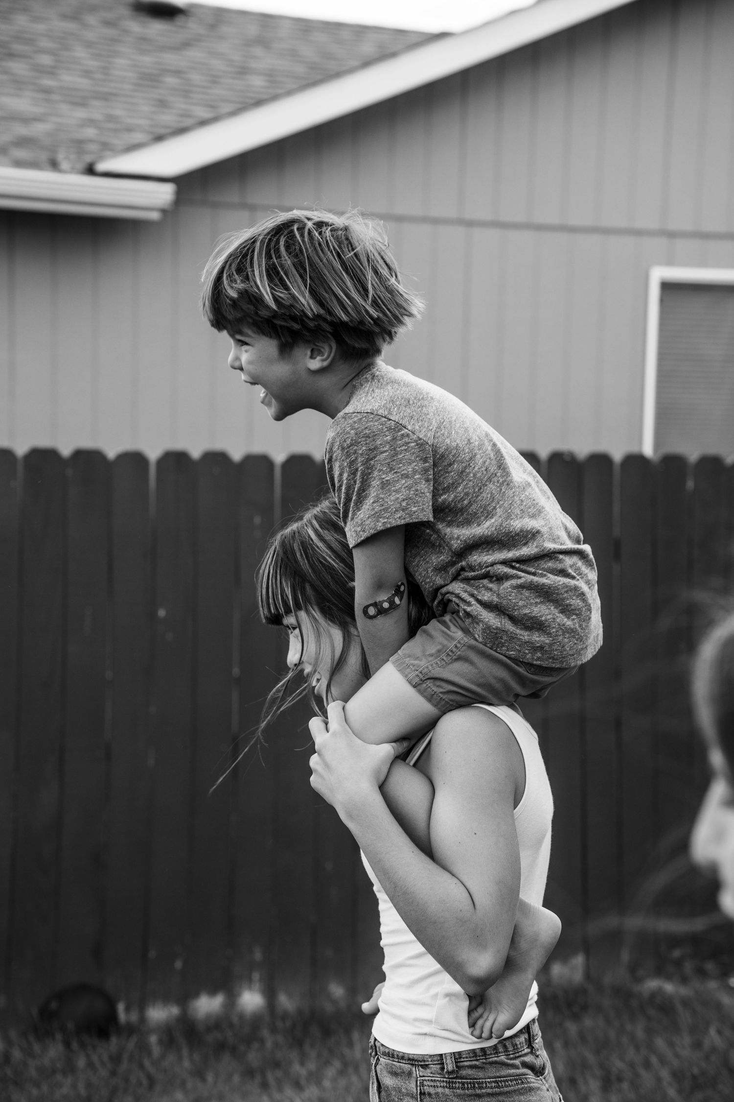 Black and white photo of a child sitting on a woman's shoulders in a backyard, laughing joyfully.