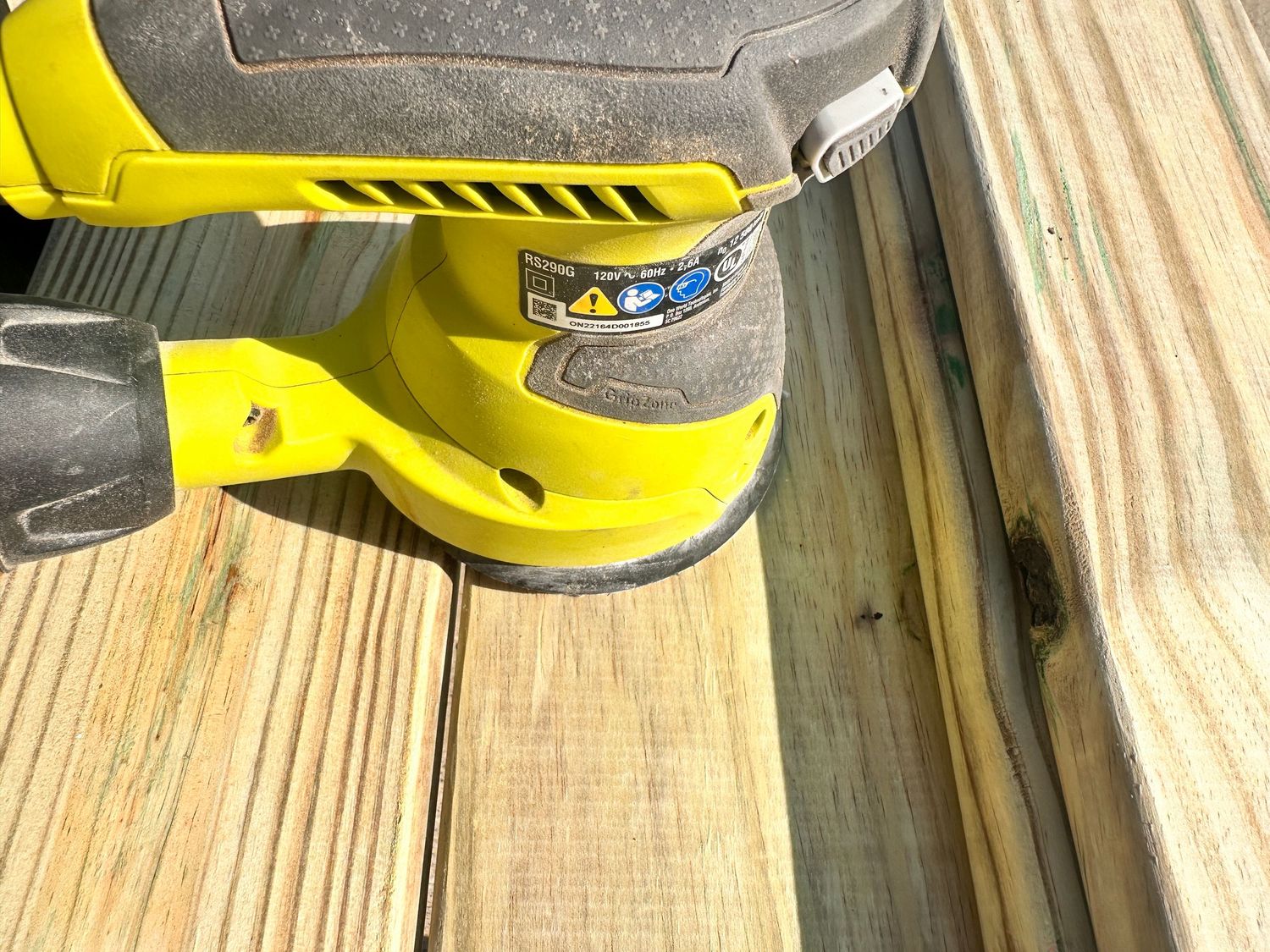 Yellow Ryobi cordless random orbital sander on weathered wood deck boards during sanding project.