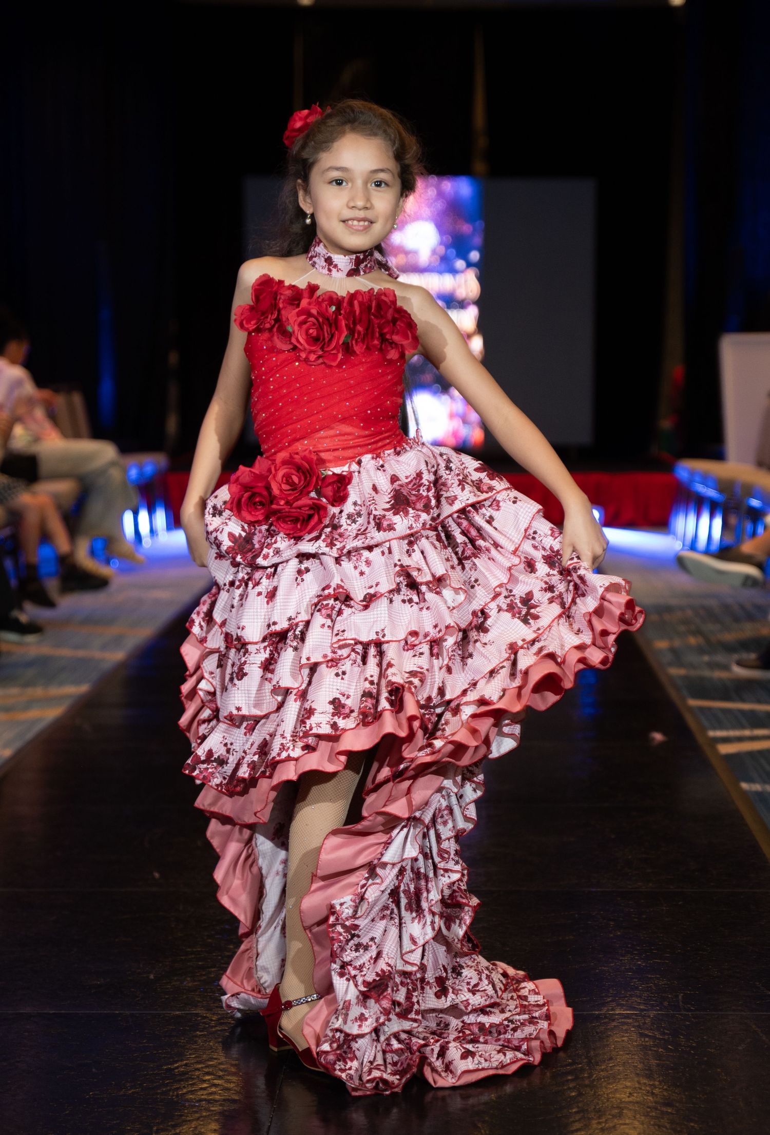 Young girl models an elegant red and pink floral ruffled gown on a fashion runway.