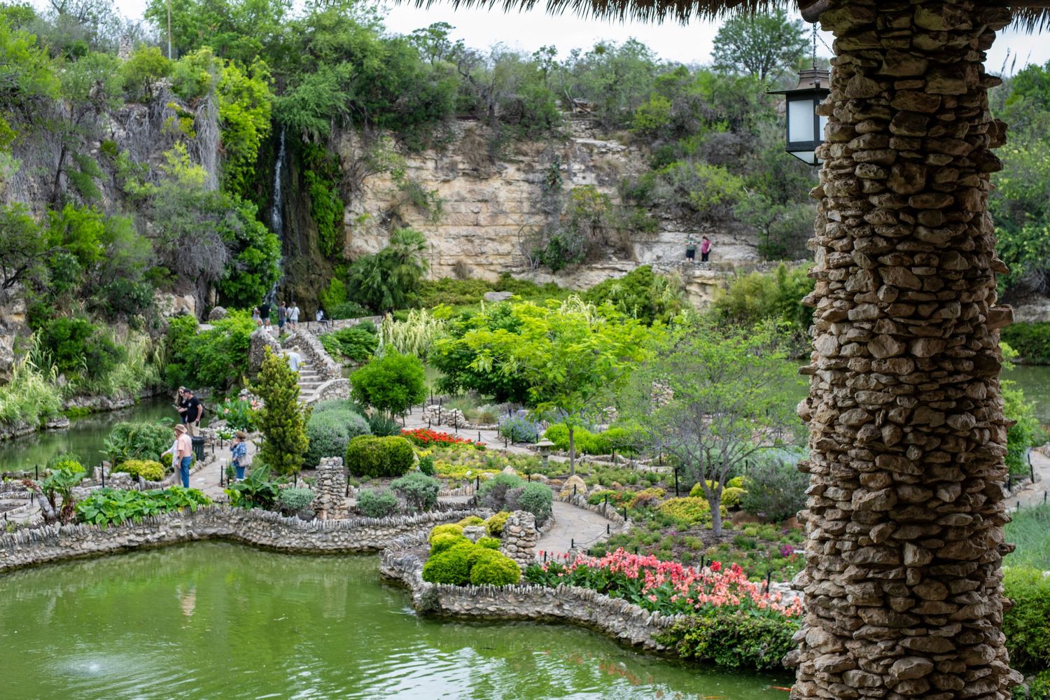 Lush botanical garden with stone walls, pond, colorful flowers, and rocky cliffs surrounded by green trees.