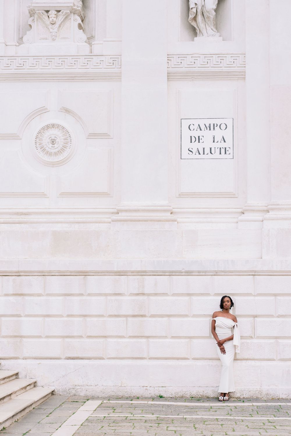 A Black woman in a white off-shoulder outfit stands against the white marble wall of Campo de la Salute in Venice.