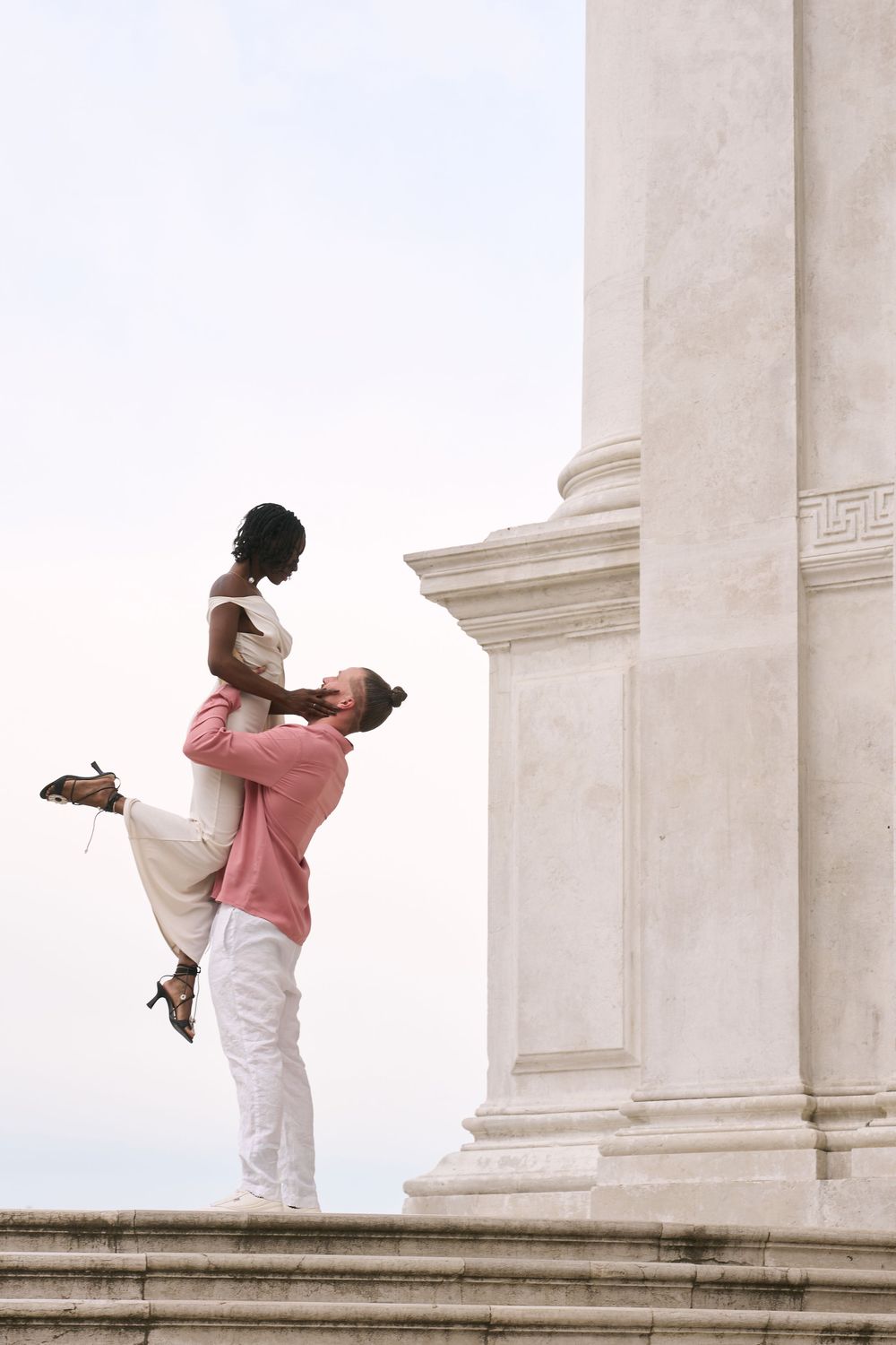 A man lifts a woman joyfully near a marble column, both dressed elegantly in white and pink attire.