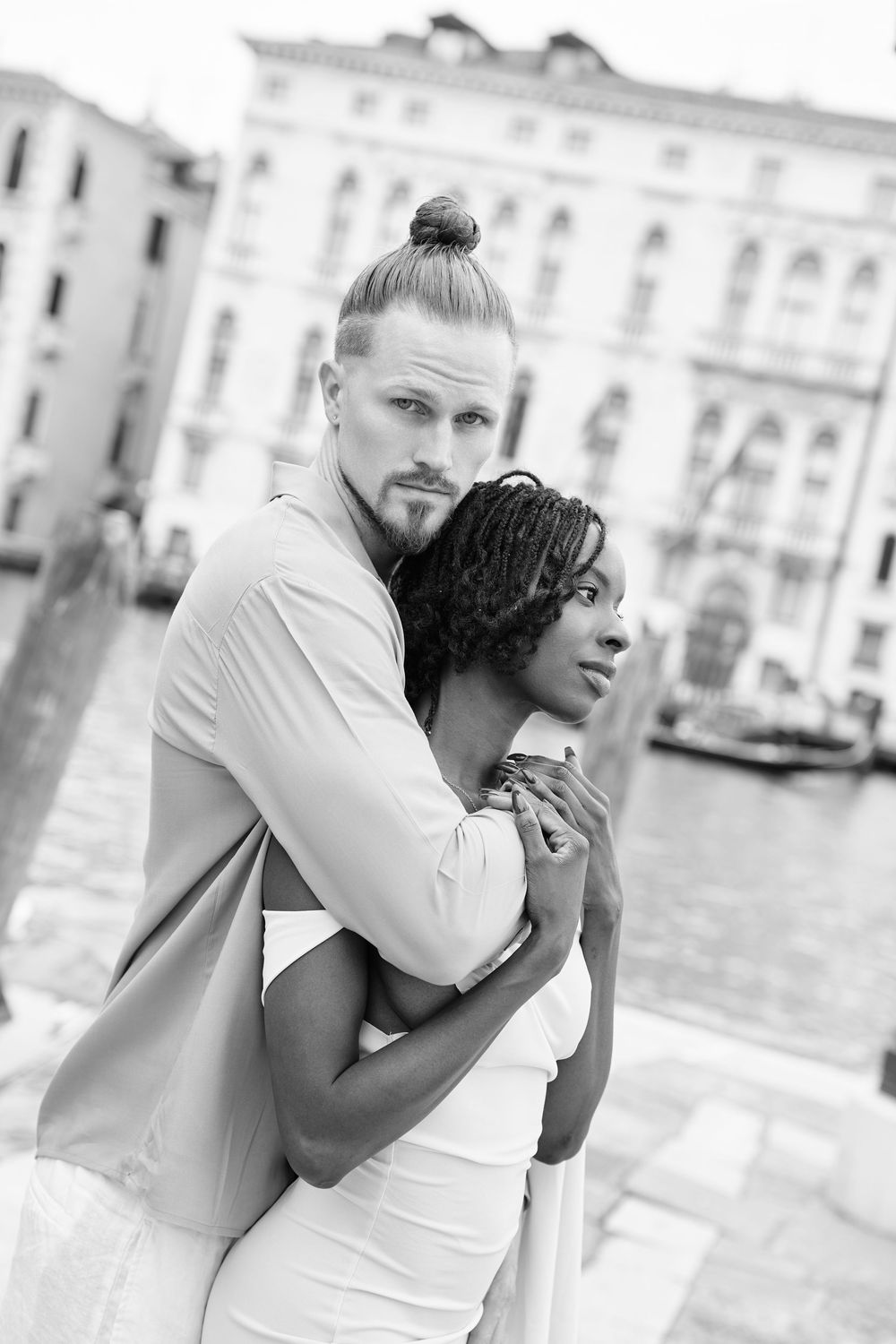 Black and white photo of an interracial couple embracing romantically in Venice, Italy, with historic buildings and canal in background.