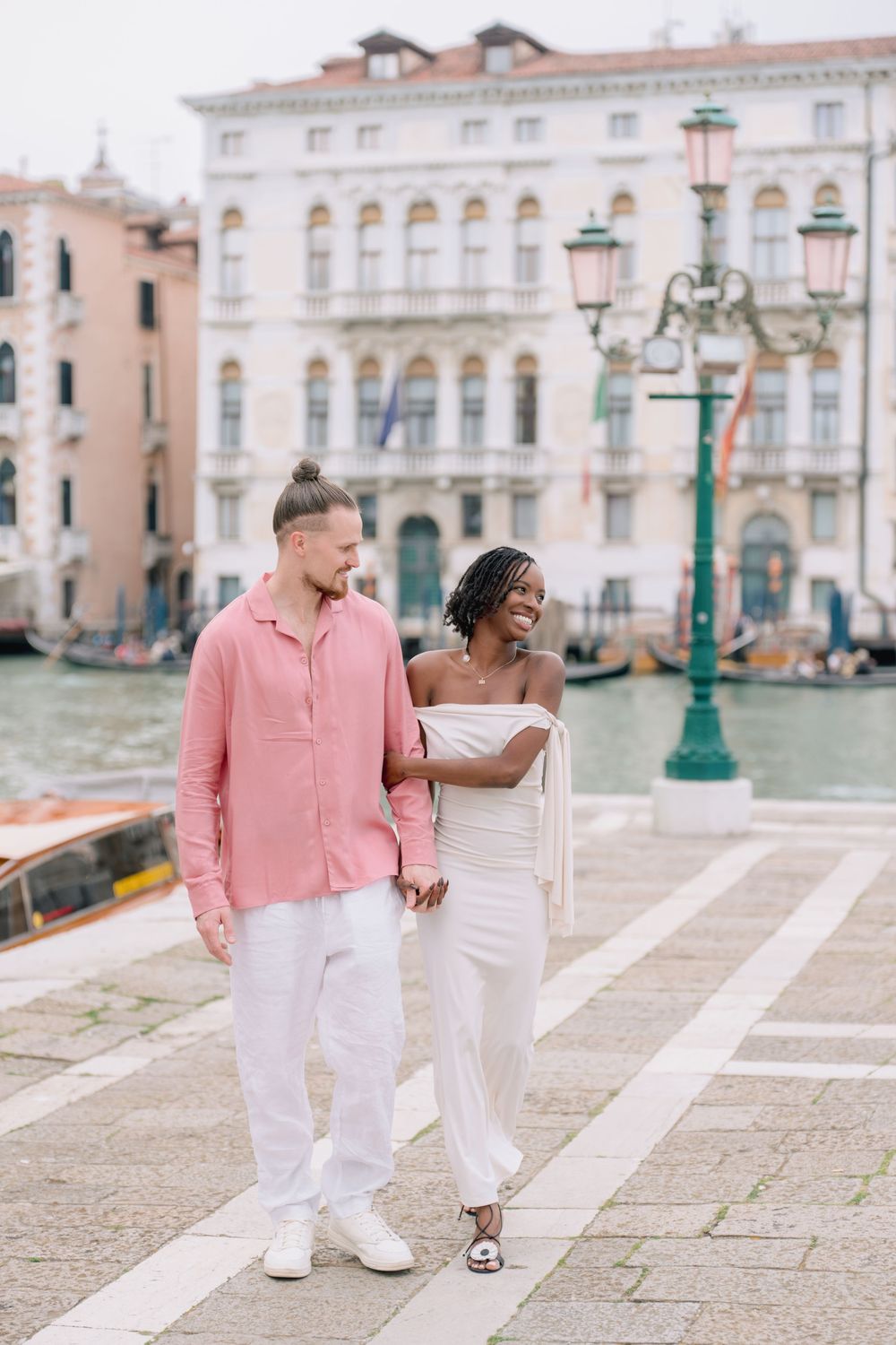 Interracial couple walking hand in hand along Venice Grand Canal waterfront, man in pink shirt, woman in white outfit.