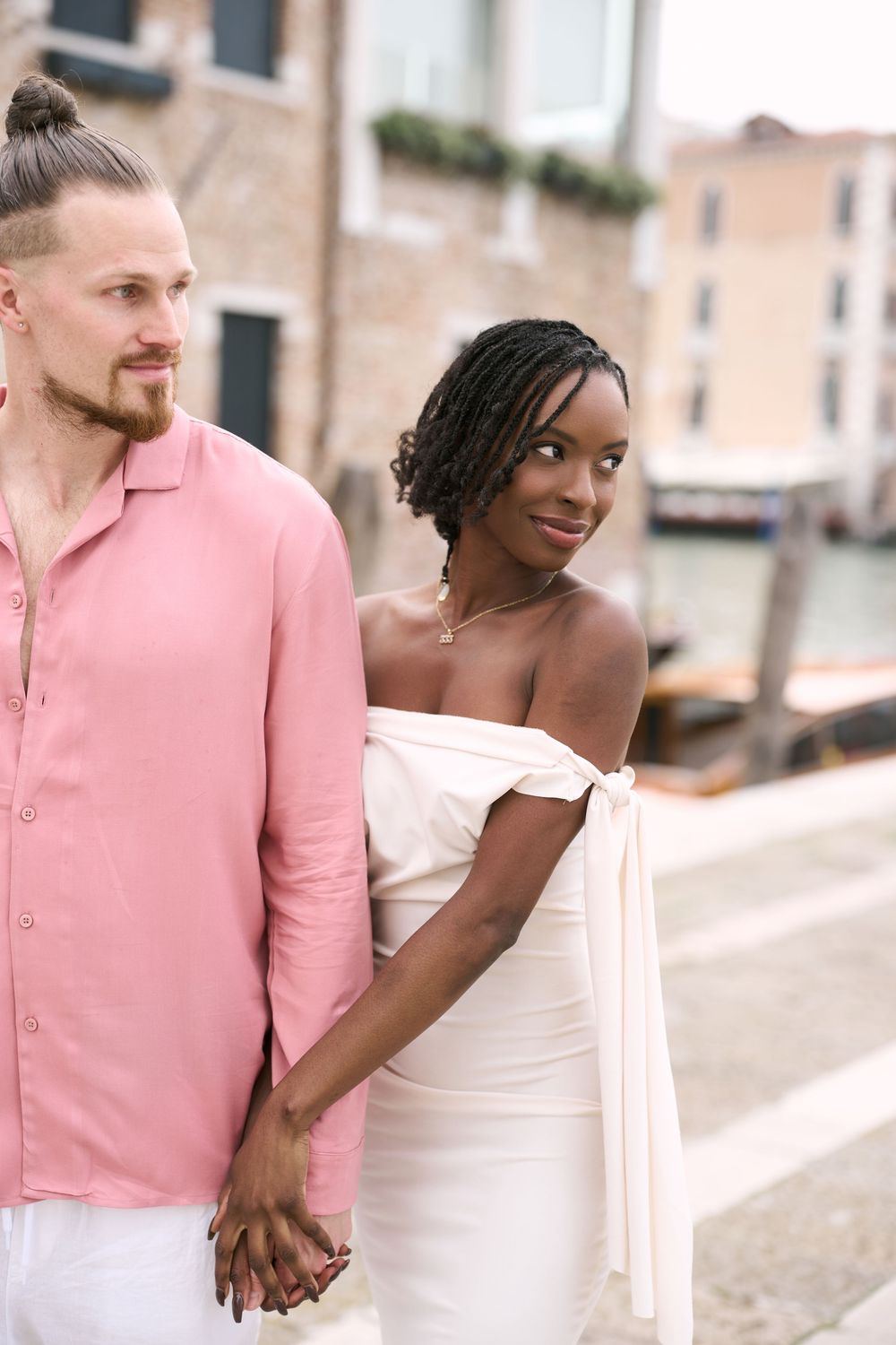 Interracial couple holding hands in Venice, woman in white off-shoulder dress smiling, man in pink shirt by canal.