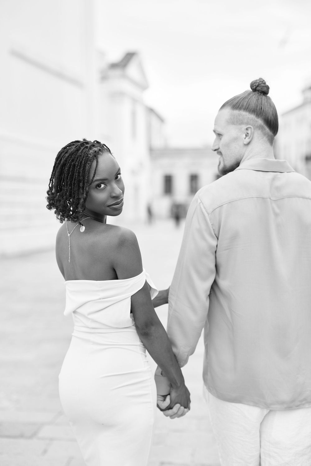 Black and white photo of an interracial couple holding hands, woman in white off-shoulder dress looks back at camera.