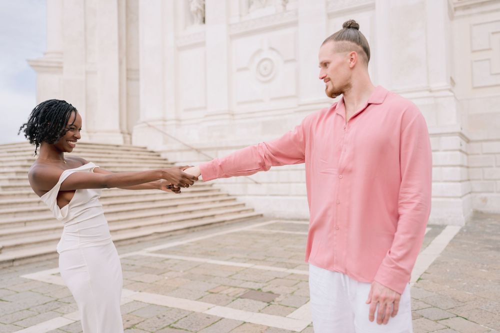 A joyful interracial couple holds hands outdoors near a white building, dressed in elegant white and pink outfits.