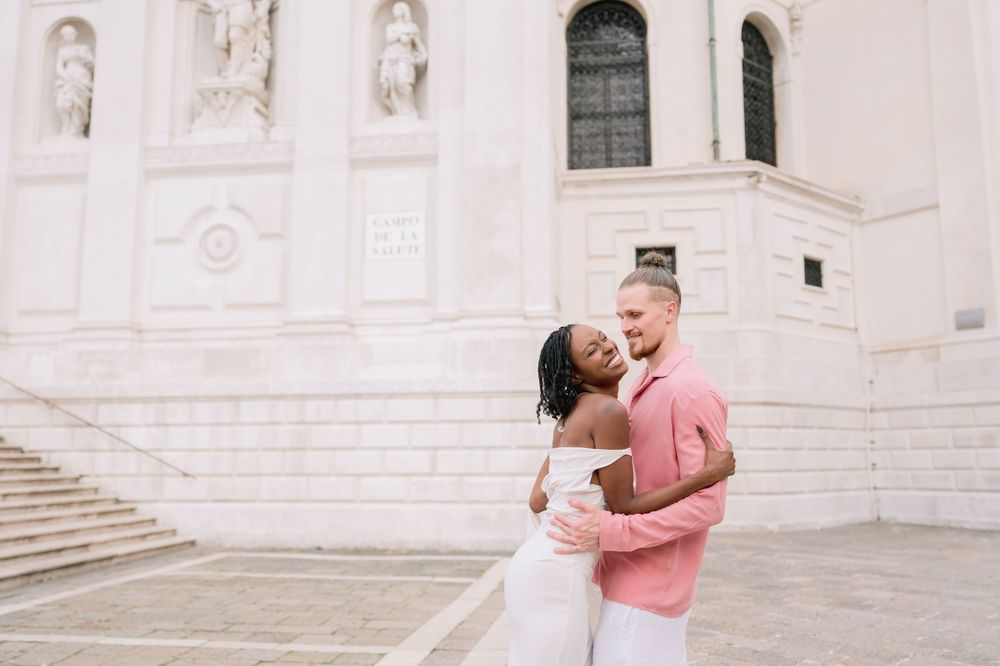 Interracial couple embracing and smiling in front of a white classical building, woman in white dress, man in pink shirt.