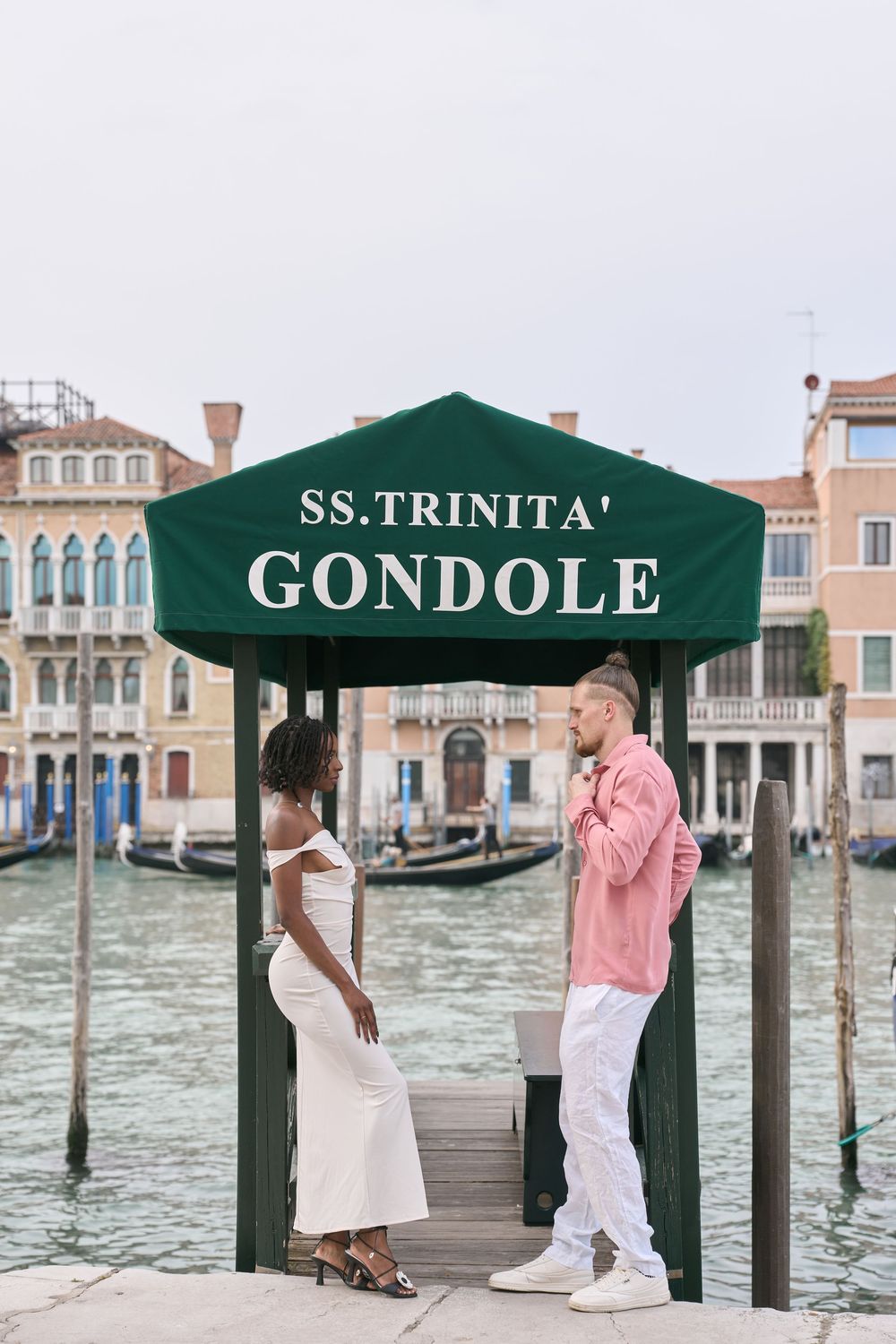 Couple standing at SS. Trinita Gondole dock in Venice, Italy, with Grand Canal and historic buildings behind.