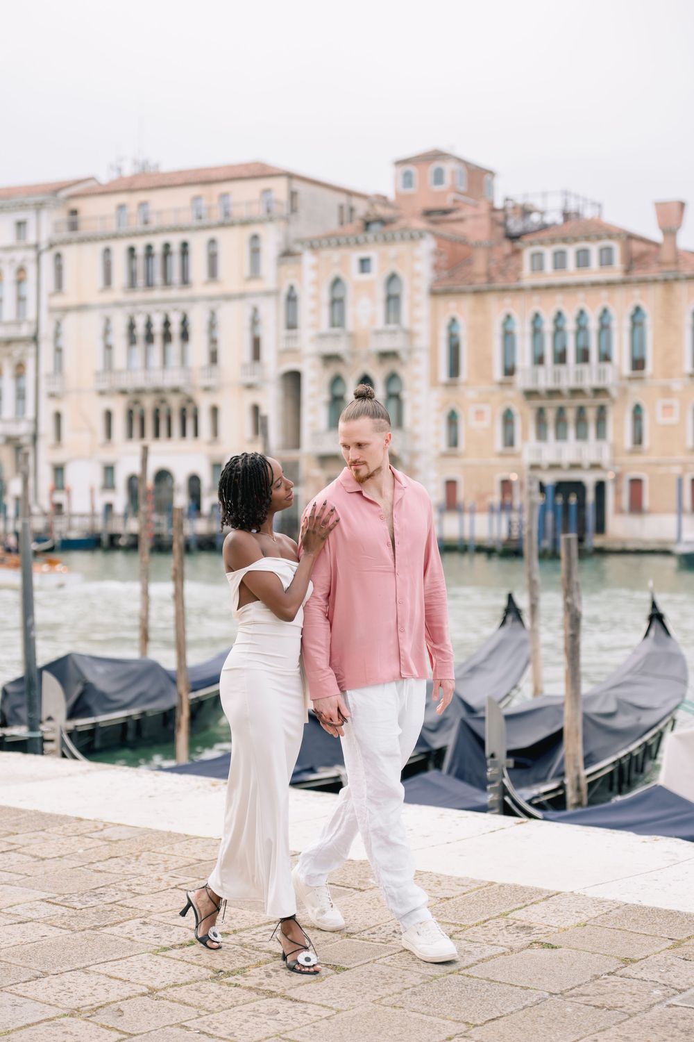 Interracial couple walking along Venice Grand Canal with gondolas and historic buildings in background.