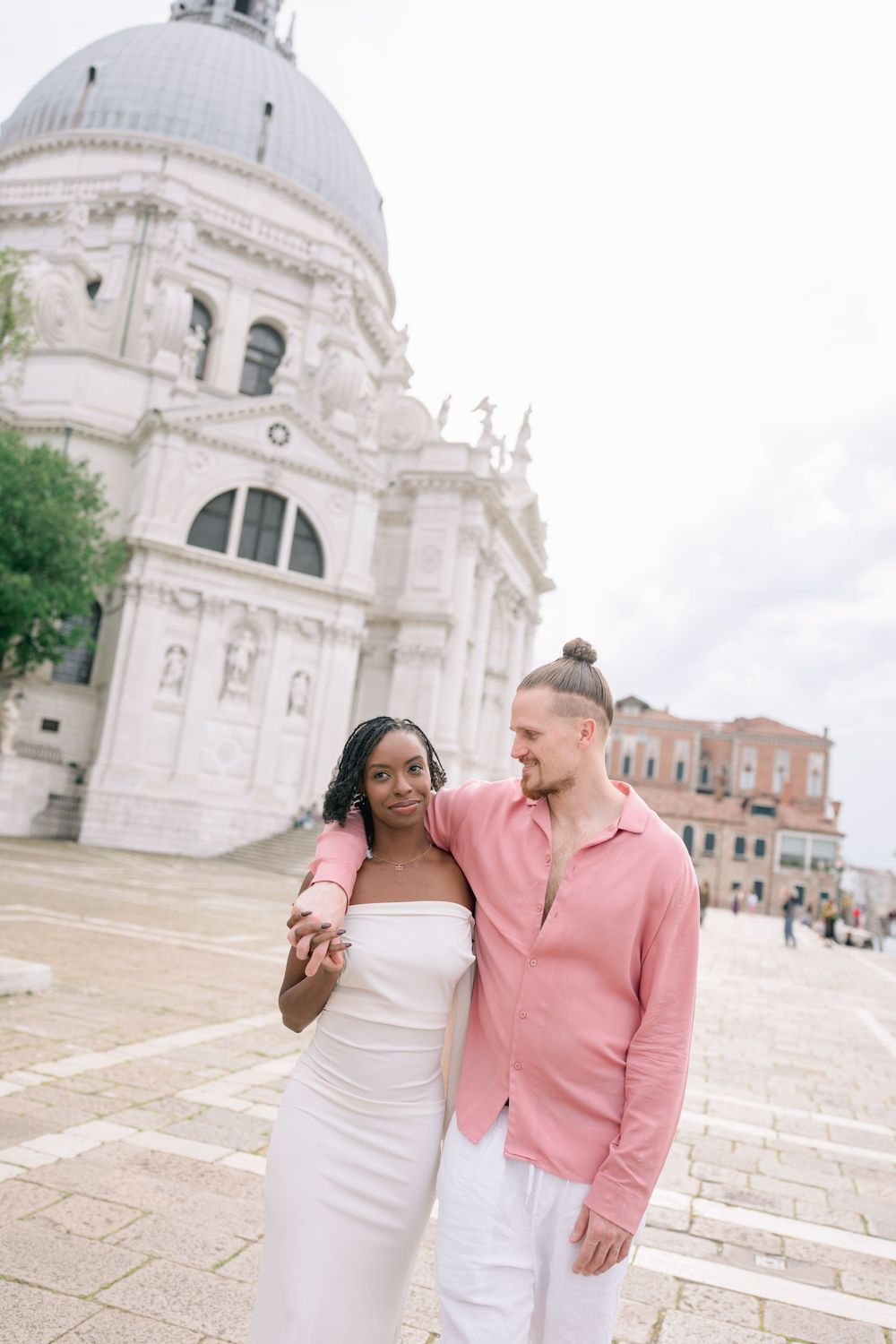 Couple posing in front of Santa Maria della Salute basilica in Venice, Italy, woman in white dress, man in pink shirt.