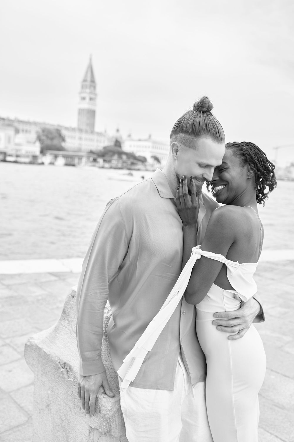 Black and white photo of a smiling interracial couple embracing near Venice waterfront with St Marks Campanile in background.