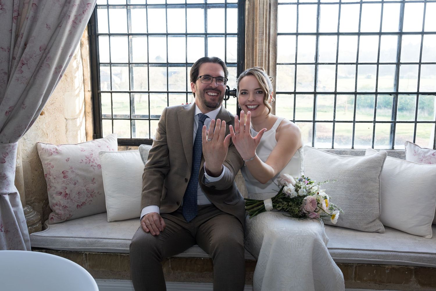 Newlyweds sitting by a window showing off their wedding rings, bride holding a bouquet in a rustic venue.