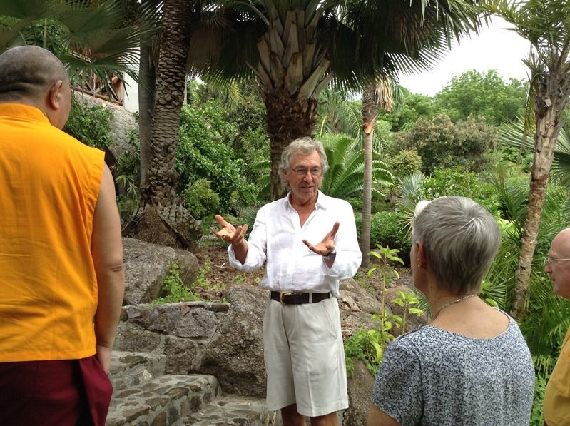 Man in white linen outfit gestures while speaking outdoors to group near tropical garden with palm trees.