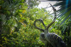Dinosaur sculpture surrounded by lush tropical greenery and palm trees in an outdoor garden setting.