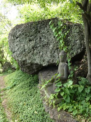 Stone Buddha statue nestled beneath a large moss-covered boulder surrounded by lush green foliage in a serene garden.
